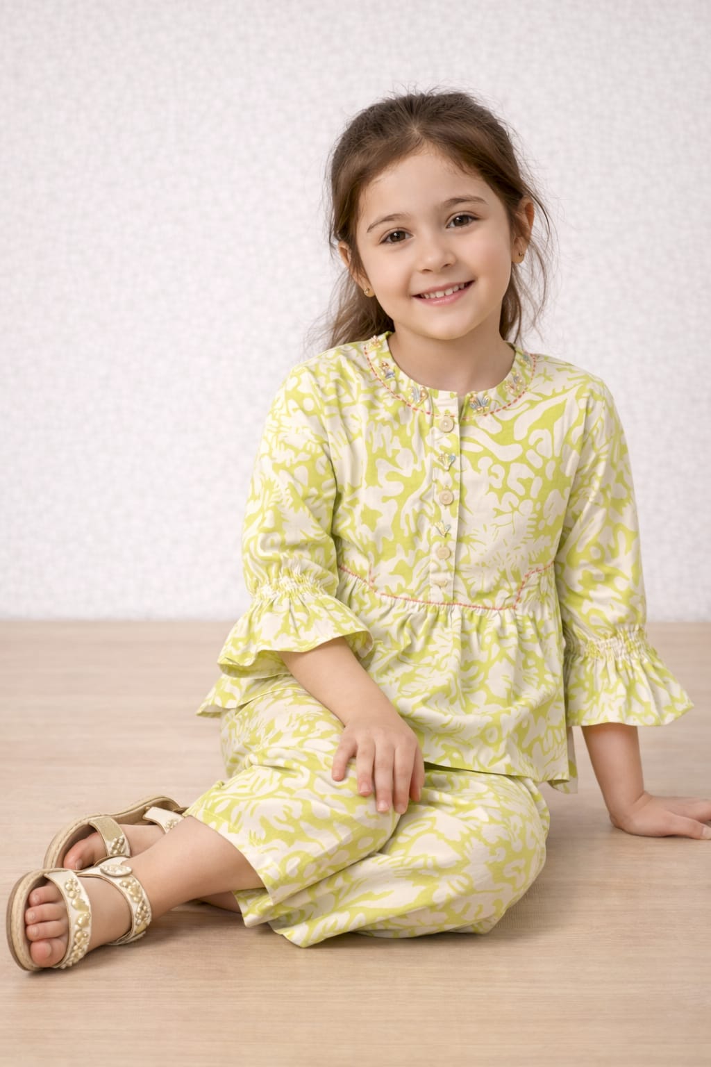 Young girl wearing a yellow floral outfit sitting on a wooden floor.